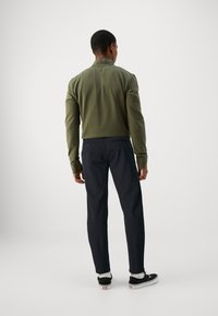 Olive green sweatshirt with a high collar, paired with dark navy trousers. The model wears black sneakers with white soles and ankle socks.