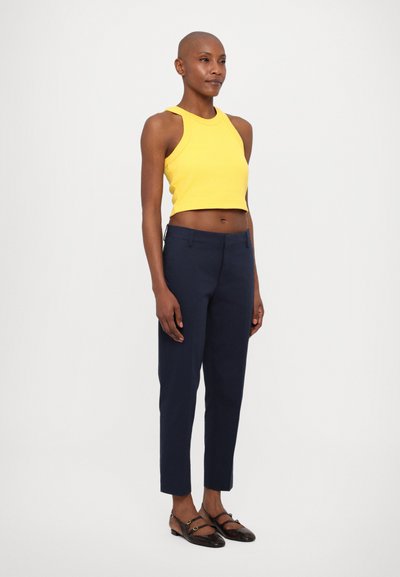 Žlutý žebrovaný crop top, námořnické přizpůsobené kalhoty a tmavé ploché boty se pásky. Hladké textury a přiléhavý design, postranní pohled.