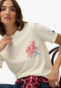 Crèmekleurig t-shirt met een rood gevleugeld leeuwgrafiek en blauwe tekst "Superdry". Korte mouwen en een casual pasvorm, met een geruite blouse gestrikt om de taille.