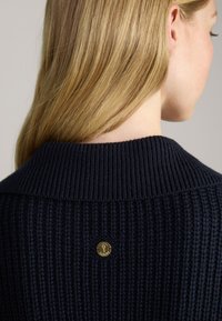 Granatowy sweter robiony na drutach z wyraźną teksturą w prążki oraz widocznym złotym logo na tylnej części kołnierza.