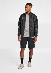 Svart lättviktsjacka med hel dragkedja, långa ärmar och mesh-accenter, bärs över en grå t-shirt och svarta shorts. Utrustad med grå sportskor.