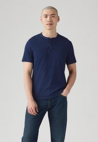 Chemise henley bleu marine à manches courtes en tissu doux, avec un col rond, une patte de boutonnage à trois boutons et une coupe décontractée. Portée avec un jean foncé.