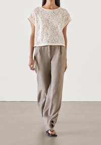 Haut court en dentelle blanche à motif géométrique, associé à un pantalon ample en lin brun clair et des tongs noires. Design simple, sans ornements.