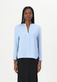 Blusa a maniche lunghe azzurro chiaro con scollatura a V e vestibilità comoda, realizzata in tessuto morbido, indossata sopra una gonna nera.