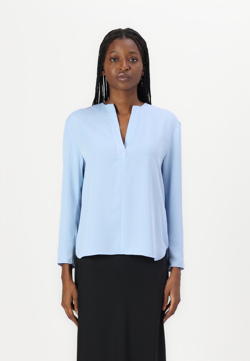 Blusa a maniche lunghe azzurro chiaro con scollatura a V e vestibilità comoda, realizzata in tessuto morbido, indossata sopra una gonna nera.