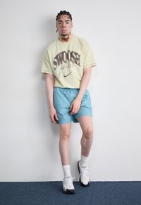 T-shirt gráfica amarelo claro com impressão castanha, combinada com calções cor teal. Ténis brancos com detalhes verdes e meias brancas. Fundo simples.