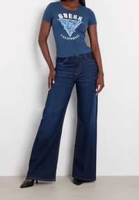 Blå bomuld T-shirt med "GUESS CALIFORNIA" grafik, kombineret med mørkeblå viddebenede denimjeans. Sorte stilethæle fuldender outfittet.