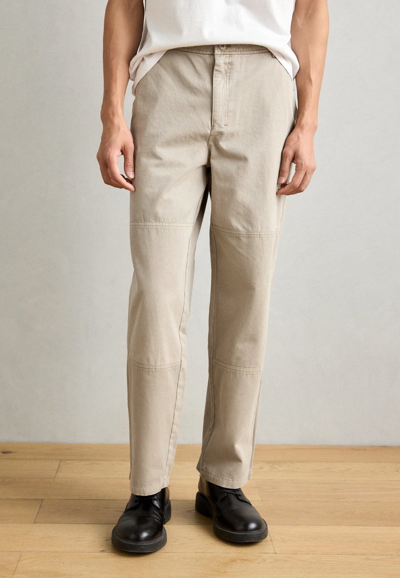 Beige cotton trousers with a straight fit, featuring side pockets, subtle stitching details, and worn with black shoes on a wooden floor.