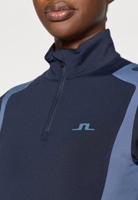 Jūras spēku zip-up puloveris ar augstu apkakli. Iezīmējas ar gaiši ziliem akcentiem plecos un mazu logo krūtīs. Gluda tekstūra.