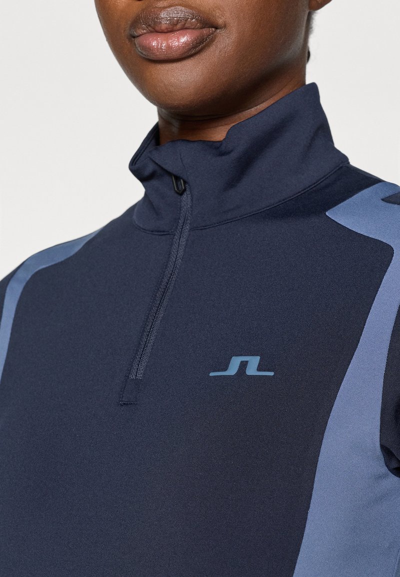 Pull en zip navy avec un col montant. Présente des accents bleu clair sur les épaules et un petit logo sur la poitrine. Texture lisse.
