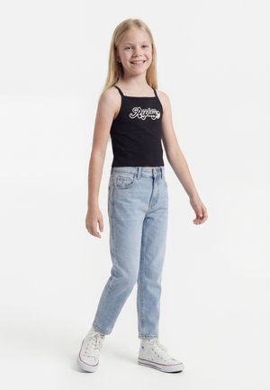 Κορίτσι που χαμογελάει με μακριά ξανθά μαλλιά, φοράει μαύρο τοπ, ανοιχτό μπλε τζιν και λευκά sneakers με ψηλή σόλα, στέκεται σε λευκό φόντο.