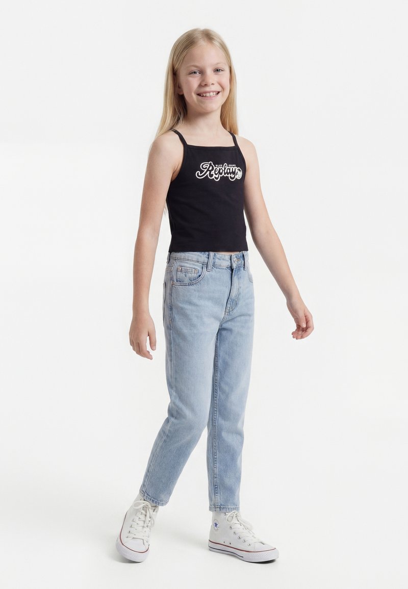 Κορίτσι που χαμογελάει με μακριά ξανθά μαλλιά, φοράει μαύρο τοπ, ανοιχτό μπλε τζιν και λευκά sneakers με ψηλή σόλα, στέκεται σε λευκό φόντο.