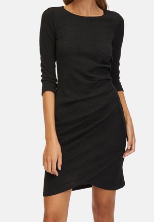 Žena oblečená v přiléhavých černých texturovaných šatech s tříčtvrtečními rukávy a asymetrickým lemem, stojící před jednoduchým pozadím.