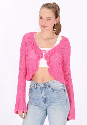 Jonge vrouw met blond paardenstaart draagt een roze gebreid kort vest over een wit cropped topje en lichtblauwe hooggetailleerde jeans, gecombineerd met stervormige oorbellen.