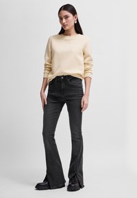 Suéter texturizado amarillo claro con un patrón de espiga, combinado con pantalones vaqueros de cintura alta negros de corte acampanado con dobladillos divididos y botas negras.