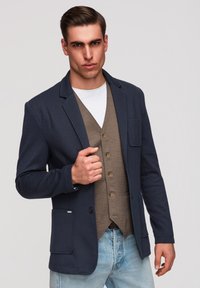 Mann trägt marineblauen Blazer, braune karierte Weste, weißes Hemd und hellblaue Jeans, posiert mit einer Hand am Revers des Blazers.