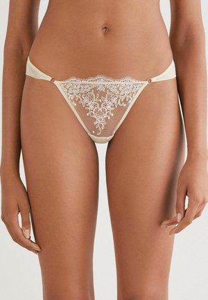 String beige transparent avec broderies florales, détails complexes, ceinture en satin et petits anneaux en métal accentuant les côtés.