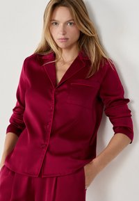 Burgundowa satynowa koszulka piżamowa z krótkimi rękawami, z kołnierzykiem z wykładanym brzegiem, przednią kieszonką i kontrastowymi lamówkami. Jedwabista faktura.