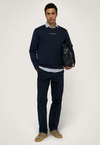 Felpa navy con logo "TRUSSARDI", indossata sopra una camicia azzurra a collo. Pantaloni scuri e sneaker olive completano l'outfit, con una borsa nera in mano.