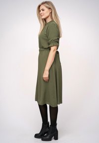 Rochie midi verde măslinie cu mâneci scurte și puff, talie legată și buzunare laterale, accesorizată cu cizme negre până la gleznă și colanți negri.