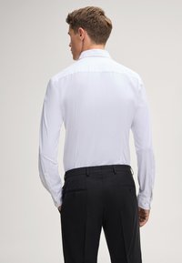Weißes Langarmhemd mit Standardkragen und geknöpften Manschetten, getragen mit dunklen Hosen, von hinten betrachtet. Gleiche Stoffoberfläche.