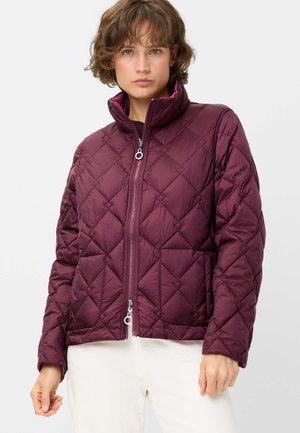 Woman wearing a quilted plum jacket with front zipper and high collar, paired with white pants, against a plain background.