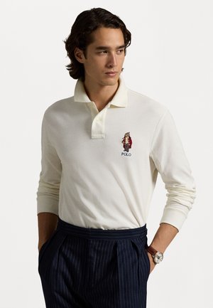 Camisa polo de manga larga en algodón crema con cuello acanalado; presenta un logo bordado en el pecho; se lleva con pantalones de rayas marinero y un reloj.