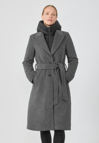 Palton lung din lână gri cu o curea, rever mari și două buzunare frontale, suprapus peste o jachetă puffer neagră cu guler înalt.