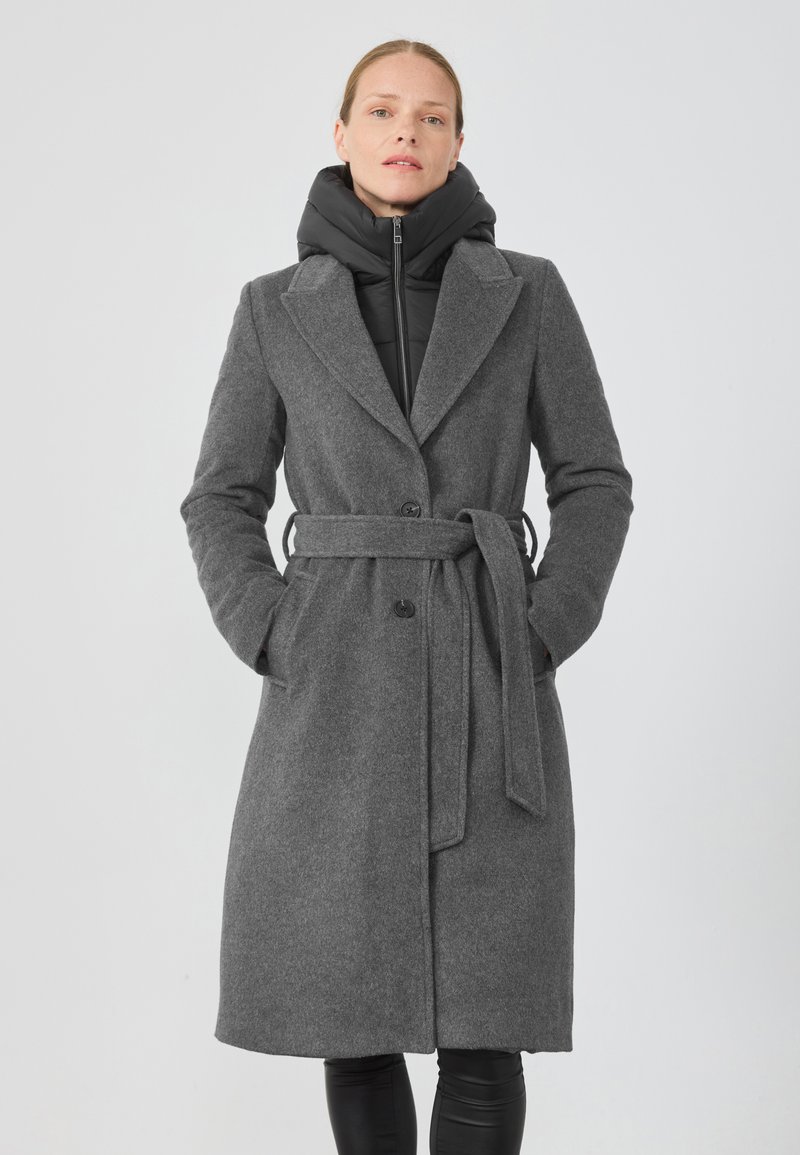 Palton lung din lână gri cu o curea, rever mari și două buzunare frontale, suprapus peste o jachetă puffer neagră cu guler înalt.