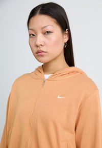 Oransje hettejakke med glidelås laget av mykt stoff, med en hvit Nike-logo på venstre bryst og hette, med en glatt tekstur.