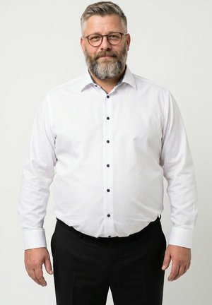 Homme d'âge moyen avec une barbe grise et des lunettes, portant une chemise blanche boutonnée et un pantalon noir, debout devant un fond clair uni.
