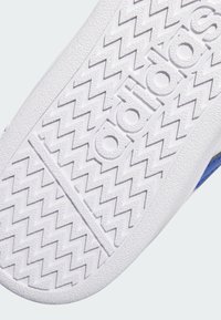 Suela de zapatilla deportiva blanca con superficies texturizadas y un patrón de agarre en zigzag; presenta la palabra "adidas" embossed en el centro.