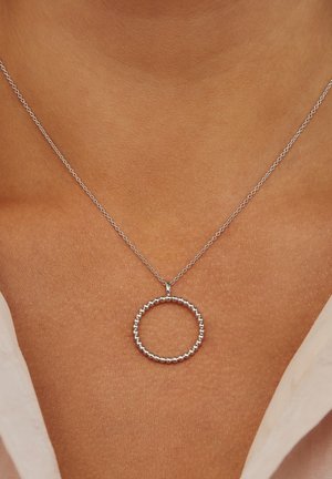 Collier en argent avec un pendentif circulaire à texture perlée, suspendu à une chaîne délicate. Gros plan sur la texture de la peau et les détails du bijou.