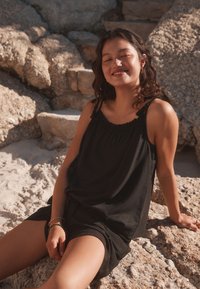 Black sleeveless top with gathered neckline and loose fit, paired with black shorts, worn by a person sitting on rocky terrain.
