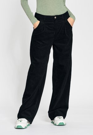 Personne portant un pantalon en velours côtelé noir ample, un haut à manches longues vert, des baskets blanches avec des semelles vertes, les mains dans les poches, debout sur un sol blanc.