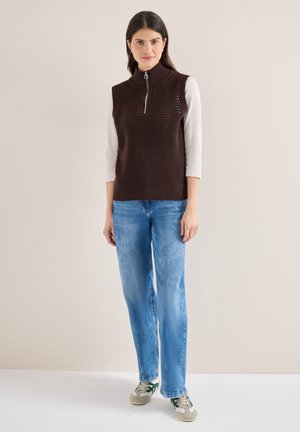 Femme debout portant un gilet marron en tricot sans manches avec fermeture éclair, un haut blanc à manches longues, un jean bleu et des baskets gris-vert sur un fond uni.