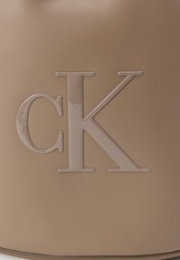 Šviesiai rudas rankinės rankinis su tekstūruotu paviršiumi, kuriame iškaltas blizgus "CK" logotipas šviesiai rudos spalvos. Paprasta cilindro forma ir minkšta konstrukcija.