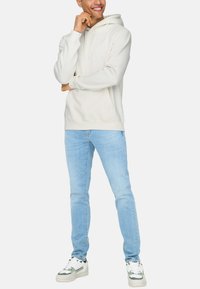 Crème hoodie met een kangoeroezak en een capuchon met trekkoord, gecombineerd met lichtblauwe jeans en witte sneakers met groene accenten.