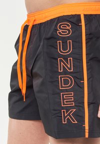 Pantaloni da bagno neri con un laccio arancione e una striscia verticale a contrasto. Presentano una grande scritta "SUNDEK" in ricamo arancione lungo il lato.