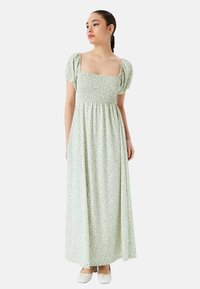 Lys grøn blomster maxi kjole med firkantet hals, smocket liv og pufærmer, der har en svævende nederdel og blød stoftekstur.
