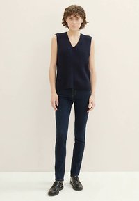 Tmavomodrý pletený top bez rukávov s výstrihom do V, s rebrovanou textúrou a rovný strih, v kombinácii s tmavomodrými úzkymi džínami a čiernymi topánkami.