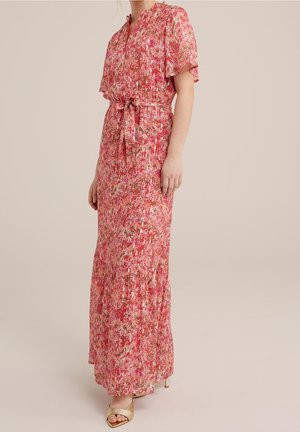 Frau trägt ein fließendes, knöchellanges rotes Blumenkleid mit kurzen Ärmeln, Taillenband und goldenen offenen Sandalen, steht vor einem neutralen Hintergrund.