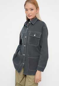 Veste en denim gris foncé avec des coutures claires, poches poitrine et fermetures à boutons-pression. Design décontracté avec une coupe confortable.