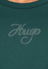 Tmavě zelená žebrovaná mikina s lesklým stříbrným logem "Hugo" umístěným uprostřed na hrudi. Kulatý výstřih, volný střih.
