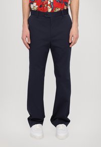 Pantalones azul marino de tela, con un diseño de pierna recta, cinturilla con pasadores y cierre de botón en la parte delantera. Combinados con zapatillas blancas.