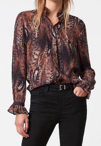 Flowy blouse with a feather pattern in earthy tones of brown and orange, featuring a band collar and ruffled cuffs. Worn with black pants.