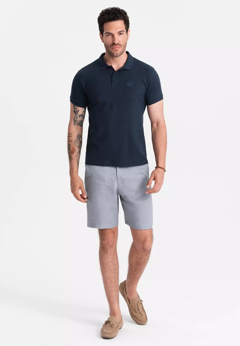Camisa polo navy de manga corta, pantalones cortos gris claro y mocasines color marrón. Presenta un sutil logo bordado en la camisa. Diseño minimalista.