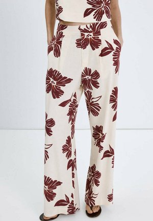 Pantalones anchos crema con estampado floral en rojo oscuro, combinados con top corto a juego y sandalias negras, modelo de pie con las manos en los bolsillos.