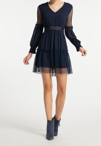 Robe bleu marine avec un décolleté en V, manches longues en résille transparente, accent en dentelle à la taille et jupe superposée à ourlet volanté. Associée à des bottines bleues.