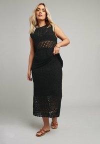 Svart virkad klänning med geometriska mönster, ärmlös design och en figurnära silhuett. Har en midjelång fåll och en mjuk textur.